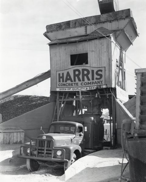 An International LF-190 truck owned by the Harris Concrete Company parked under a concrete chute. There is a man in the driver's seat. The original caption reads: "That construction work is buzzing right along in Corpus Christi, Texas, is well evidenced by the fact that the Harris Concrete Company has had to make a sizable addition to its fleet. This addition consists of six new-type Model LF-190 six-wheel Internationals equipped with 6-1/8-yard Jaeger mixers. One of these is shown at its plant taking on a load and also delivering it to a new street job on the outskirts of the city. With these new model additions the Harris fleet consists of 30 units, 21 of which are equipped with mixers."