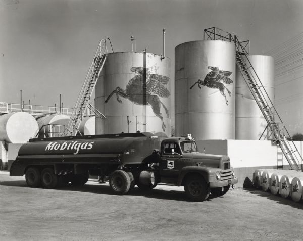 A man is driving a truck marked "Mobilgas" which is owned by the Magnolia Petroleum Company. The truck is in front of several large petroleum storage tanks. The original caption reads: "One of four L-195 International trucks purchased by the Magnolia Petroleum Company for use in making deliveries from its San Antonio plant is shown at this plant. It is shown with Trailmobile tandem trailer, the tank of which is provided with five compartments. These L-195's travel 300 miles south, 150 miles west, 60 miles east, and 90 miles north, making deliveries to dealers and Magnolia's own stations. Some 52 motor trucks are based at the San Antonio plant, 43 of which are Internationals."