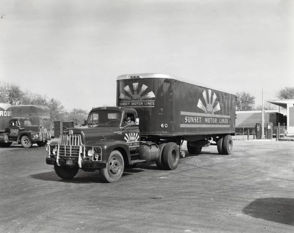 A man driving an International L-195 truck owned by Sunset Motor Lines through a parking lot. A filling station is in the background. The original caption reads: "These photographs wre taken of a Model L-195 International operated by the Sunset Motor Lines, whose headquarters are in San Angelo, Texas, at their San Antonio branch. The truck has a wheelbase of 242 inches and is shown with 32-foot trailer. Some eleven tractor trucks and thirteen straight trucks are operated out of the San Antonio branch. E.A. Hunger is planning to visit San Angelo and get a full article on the big Sunset fleet which includes 170 tractor trucks and 200 trailers."