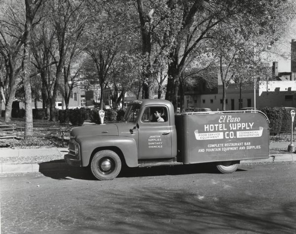 A man sitting in an International truck owned by the El Paso Hotel Supply Company along the side of the road near a park. The text on the side of the truck reads: "El Paso Hotel Supply Co.; Food Service Equipment, Commercial Refrigeration; Complete Restaurant, Bar, and Fountain Equipment and Supplies."