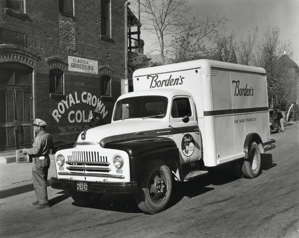 Juan B. Marquez, driver of an International L-150 truck for Borden's Fine Dairy Products, delivers a crate of dairy products to El Aguila Groceries. The building behind the truck bears the advertisement "Drink Royal Crown Cola." The original caption reads: "New L-150 International of 154-inch wheelbase, one of some 40 trucks operated by Bordens in El Paso. According to the driver, Juan B. Marquez, it travels an average of 30 miles and makes 92 stops a day. The insulated body is 12 by 7 1/2 by 6 feet in size."
