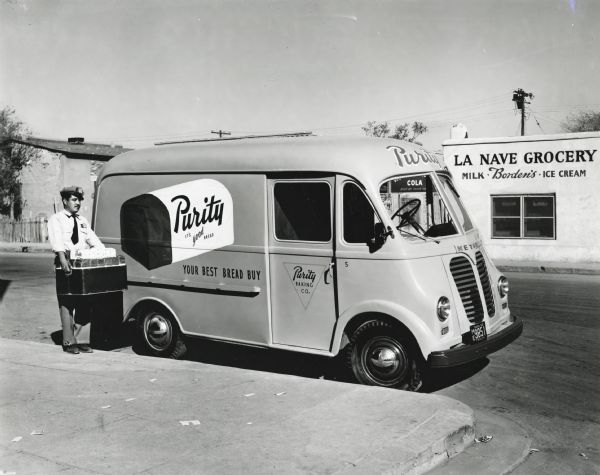 A man unloads a box from the rear of an International LM-120 with Metro body owned by the Purity Baking Company. The truck is parked on the side of a street in front of La Nave Grocery. The original caption reads: "The Purity Baking Company of El Paso, Texas, operates 14 International multi-stop delivery trucks equipped with Metro bodies. Four of these are the new LM-120 models with new all-steel Metro bodies. One of these, painted a distinctive orange with the red-and-white Purity bread loaf wrapper reproduced on the side panel in jumbo style, is shown in accompanying illustrations. The company operates 28 trucks, 23 of which are Internationals. Some of these are larger, heavy-duty trucks for longer runs north to Las Cruces, and southeast to Ft. Hancock and Esperanza. In El Paso the company operates 19 routes on which the trucks average 42 stops and 40 miles per day. The new all-steel Metro body shown is 9 1/2 feet long and provides convenient arrangement of bread boxes in the payload compartment and of special packages on the engine cover for efficient and quick handling."