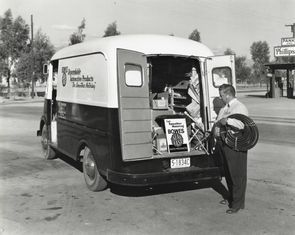 A man holds a coil of tubing while standing near the opened back doors of an International truck with Metro body owned by Bowes Auto Products. The text on the side of the truck reads: "Bowes Dependable Automotive Products; For Smoother Motoring," "Seal-Fast," and "C.C Samuel, Dist.; C.R. Carlisle, Salesman." A Phillips filling station is across the road in the background.