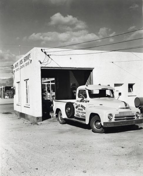 An International L-110 truck owned by White Brothers Service automotive repair company parked near the opened door of a garage building. The original caption reads: "White Brothers of Tucson, Arizona, are relying on their recently purchased L-110 International with 8-foot pickup body as an important adjunct in giving their customers the best of automotive service station. It is painted an attractive Palomino (No.102) yellow with red-shaded black lettering. The truck has a wheelbase of 127 inches."