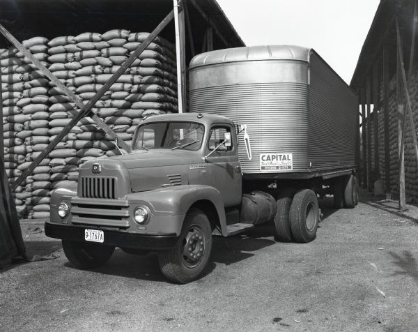 An International L-195 truck with a Fruehauf trailer drives through an outdoor storage area at Capital Feed & Seed Company. The original caption reads: "The Capital Feed & Seed Company, 312 South Fifteenth Avenue, Phoenix, Arizona, utilizes the recently purchased L-195 International with 35-foot stainless steel Fruehauf trailer shown to make two weekly trips a week to Los Angeles. On each of these trips the truck with the trailer hauls 20 tons of grain and feed and travels 500 miles a day. It is shown at the company's plant in Phoenix with sacked grain in open sheds shown in the background. The truck has a wheelbase of 157 inches. The company operates fifteen trucks of which two are tractor trucks."