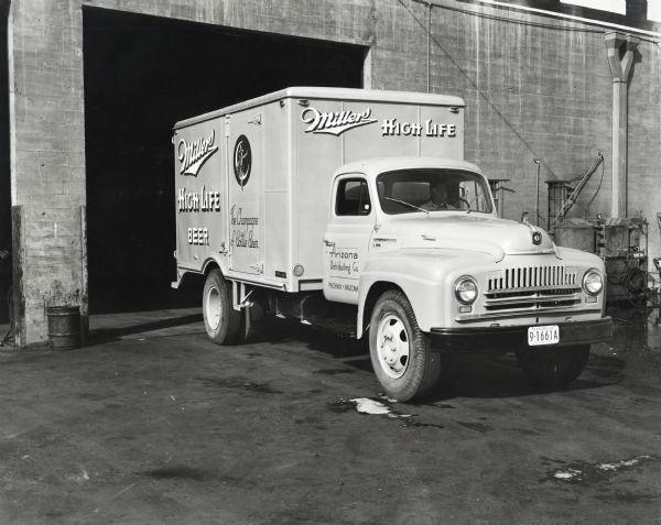 An International L-182 truck owned by the Arizona Distributing Company parked near an opened garage door of a warehouse. The truck advertises Miller High Life beer: "The Champagne of Bottle Beer." The original caption reads: "The Arizona Distributing Company of Phoenix, Arizona, has recently purchased two new Model L Internationals, one an L-162 of 154-inch wheelbase and, another, an L-182 of 172-inch wheelbase, which is shown in accompanying illustrations. The company operates a fleet of five trucks, four of which are Internationals. Besides Phoenix, it has distributing points for Millers High Life Beer at Tucson and Flagstaff. The truck is equipped with a 12-foot Fruehauf standard body and has a capacity of 440 mixed cases. C.J. Minning is manager of this distributorship."