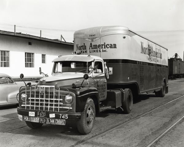 A man drives an International L-180 truck outfitted with a North American Van Lines Inc. trailer down a road next to a railroad track. Train cars, a building, and an automobile are behind the truck. The original caption reads: "These pictures show a new L-180 International of 160-inch wheelbase and a 32-foot Fruehauf van-type trailer owned by the North American Van Lines, Inc. by an owner-operator arrangement. It is used entirely in furniture moving coast to coast. The H & R Transfer & Storage Company has a fleet of twenty-four trucks, six of which are utilized in North American nation-wide service. H.J. Hart is president of the concern."