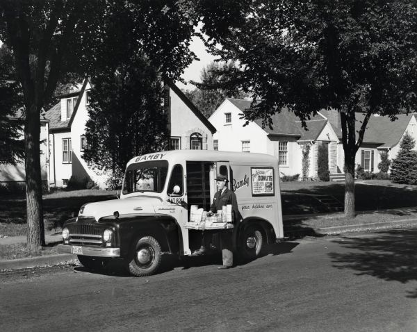 A man removes a wire tray filled with wrapped packages from an International LB-110 truck owned by the Excelsior Baking Company. The truck is parked along the curb of a residential street, and the text on the truck reads: "Bamby Delicious Donuts; Macaroon, Honey Nut, Orange, Pineapple, Raised, Bismarks; Ideal for Lunches." The original caption reads: "Twenty-five new LB-110 International trucks with special "to-the-door" delivery bodies have recently been added to the big fleet of 124 units (123 Internationals) operated by the Excelsior Baking Company of Minneapolis. One of these, painted brilliant yellow and maroon with the trade name Bamby prominently lettered thereon, is shown with driver about to make a delivery in a residential section of North Minneapolis. In keeping with the special design of this truck is the message "at your kitchen door" printed on the side of the truck. Two sets of driving controls are provided on the new LB-110 for stand-drive and sit-drive and the two brake pedals operate independently of each other. Starting and stopping without clutching or shifting is made possible by means of a new converter-coupling which automatically provides torque multiplication for starting and accelerating. In making his deliveries of rolls, cookies, cakes, doughnuts, and various kinds of bread, the driver can conveniently work from the rear or from the front as shown. Sixteen steel racks are provided for bread and a space below 12-inch high is available for miscellaneous goods.  The Excelsior Baking Company operates some 69 routes in Minneapolis and 40 in St. Paul. Besides the delivery trucks, such as shown, the company uses a number of transport trucks to convey goods from its main plant to several substations."