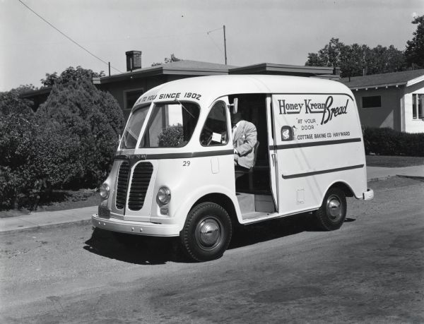 A man drives an International LM-122 truck owned by the Cottage Baking Company past several houses on a residential street. The text on the truck reads: "Honey Kream Bread, 'At Your Door,' and Oven to You Since 1902." The original caption reads: "Ed C. Friedrichs, proprietor of the Cottage Baking Company of Hayward, California, is shown in accompanying illustrations at the wheel and also at the rear proudly viewing one of five new LM-122 Internationals with Metro bodies he has purchased. He also operates thirteen KB-3-M Internationals, all being used for door-to-door delivery and sales. The front of each truck serves as a counter, so to speak, and the rear for supplies. Mr. Friedrichs says his is the only customer door-to-door delivery bakery in the Bay area. He also has a large K-5 International transport truck. The Cottage Baking Company also operates two stores. It was established in 1902 by Mr. Friedrich's father."