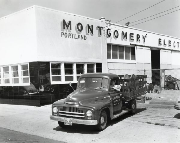 A man drives an International L-120 motor truck owned by the Montgomery Electric Company out of a fenced parking lot. The company building is behind the truck. The original caption reads: "The Montgomery Electric Company of Portland, Oregon, is the new Model L-120 International motor truck shown in accompanying illustrations. This concern is engaged in the electrical contracting business and one of its important activities is the building of transmission and distribution systems. It has done considerable work on systems for the Bonnevile Power Administration. It also has the contract for electrical work in one of the biggest apartment houses in Seattle. Its field of operations comprises the states of Oregon, Washington, and Idaho, and trucks are used to haul material from job to job in this area. The company also operates a retail store in Portland, which is shown in the background of the accompanying illustrations."