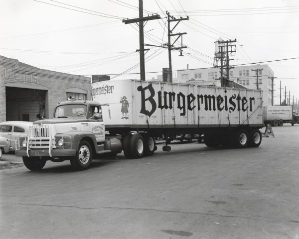 An International L-205 truck owned by the Golden Brand Products Company drives down a road in front of industrial buildings. The truck is marked: "Burgermeister." A truck is in the background reads, "Los Angeles - Seattle Motor Express." The original caption reads: "The Golden Brand Products Company of 201 Second Street, Oakland, California, distributor of San Francisco Brewing Corporation's Burgmeister Beer, recently purchased the new Model L-205 International "Roadliner" shown in accompanying illustrations. This model is one of the new L line and has a wheelbase of 157 inches. It also has a trailing axle.<p>The truck is shown with 35-foot open-top C&C trailer which has a capacity of 1,100 cases of pints and 1,900 cases of canned beer. This new model truck and also an International K-12 with trailer are used to bring in bottled and canned beer to the company's Oakland warehouse, shown in the background of the pictures, from the brewery in San Francisco. Each truck makes three trips a day. These trucks are part of a fleet of 25 units, 17 of which are Internationals, mostly K-5's and K'7's. These other trucks are called route trucks and are equipped with beverage bodies. E.M. Molakides is general manager of the Golden Brand Products Company."