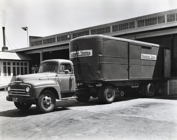 A driver backs up an International L-165 truck and trailer owned by Hagstrom's Food Stores, Inc. to a loading dock. The original caption reads, "Hagstrom's Food Stores, Inc. of 29th Street and Broadway, Oakland, California, which operates 50 stores in the Bay area, recently purchased the new-type Model L-165 International "Roadliner" truck shown with 25-foot van-type Trailmobile semi-trailer. The truck is shown at the concern's central warehouse's loading platform. It has a wheelbase of 154 inches. Both truck and trailer are painted an attractive red. Hagstrom's also have a bakery and creamery plant. They have a fleet of 31 trucks and 12 semi-trailers. They operate a total of eight Internationals (the new L-165, a K-11, two K-8's, and three K-6's), all purchased in recent years."