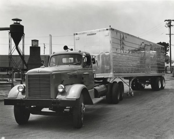 Man driving an International W-3042-L truck with a Trailmobile trailer down a dirt road. The truck and trailer were owned by the Ragusa Trucking Company. The original caption reads: "The Ragusa Trucking Company of 190 East 3rd Street, Pittsburg, California, has just purchased five big Western Internationals to be used for heavy long-distance hauling in Southern and Central California. Three of these are Model W-3042'L's equipped with Cummins diesel engine and trailing axle, and the other two are W-4064-L's with Cummins engine and dual drive. Each will be used with 35-foot flat-bed Trailmobile trailers. One of the diesel W-3042-L's is shown before delivery at the Trailmobile plant in Oakland hitched to a van-type trailer. All these trucks are equipped with the new Comfo-Vision cabs."
