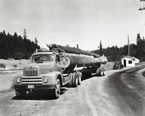 An International L-210 truck hauls a Peerless trailer loaded with Hemlock logs. The truck is parked along the side of a road with a wooded area, automobile, and small buildings in the background. The original caption reads: "The new Model L-210 International shown in accompanying illustrations is operated by Glenn E. Park of Estacada, Oregon, logging trucker. Mr. Park also operates a K-11 International with single drive and K-8-F with dual drive. The L-210 shown is equipped with trailing axle and has a wheelbase of 193 inches. The truck is shown pulling a Peerless trailer. The load consisted of 5,500 feet of Hemlock logs, which were being hauled 55 miles from Ladee Flat to paper mill in Oregon City."