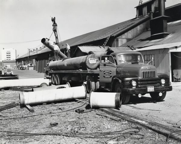 Two men standing on a pipe on the bed of a International truck use a crane to unload one of the pipes. A long building, possibly a warehouse, is in the background. Other pipes lay on the ground near the truck. The original caption reads: "The northwest division of the American Pipe & Construction Company makes use of two big International motor trucks with 35-foot flat-bed trailers to transport heavy concrete and steel pipe, plates for steel elevated tanks, and similar products to various jobs in Oregon, Washington, Idaho, and Montana. One of these trucks, a new Model L-195 with 157-inch wheelbase International Super Red Diamond Engine and Comfo-Vision cab, is shown being loaded at the company's big Portland plant.
The two lengths of concrete pipe shown were part of an order for an outfall sewer for the city of Olympia. Each length was 32 feed long, 30 inches in diameter inside, and weighed 8,100 pounds. The steel water pipe shown being loaded with the two concrete lengths was 20 inches in diameter, was coated inside with coal tar enamel and outside with asbestos paper, and was for a new water system for the Tacoma-Seattle airport."