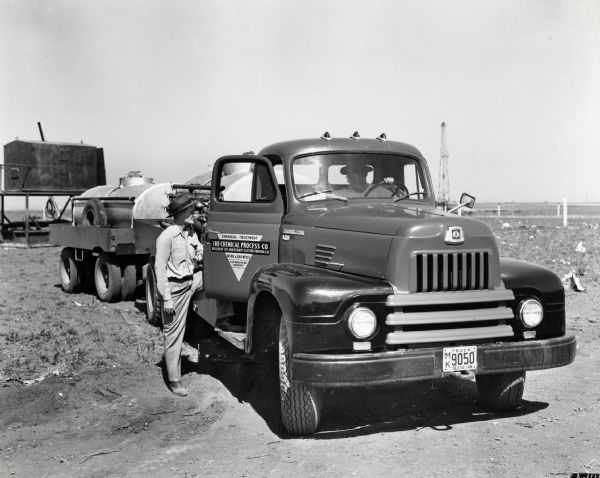 A man opens the passenger door of an International truck owned by the Chemical Process Company to speak to the truck's driver.  The automobile hauls a trailer and is parked in a dirt field; The text on the vehicle's door reads: "The Chemical Process Co.; Chemical Treatment of Oil & Gas Wells; Experienced Service."