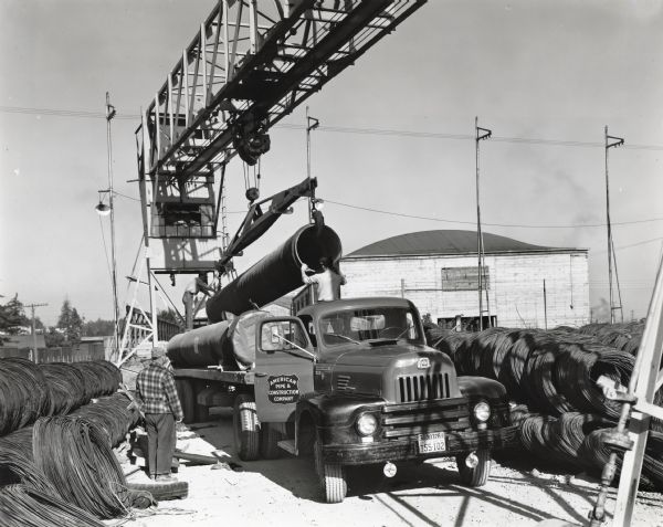 Men guiding a pipe onto the back of an International truck bed as it is lowered with an overhead crane. The truck, owned by American Pipe & Construction Company, is parked in what appears to be an storage area, surrounded by coils of wire.