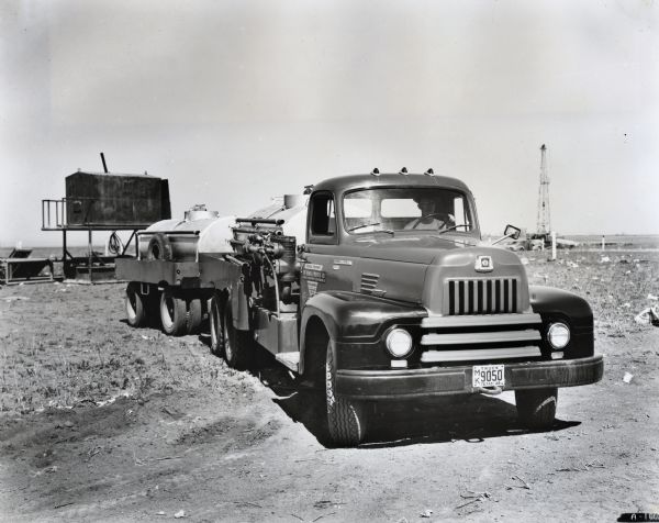 A man drives an International LF-192 truck owned by the Chemical Process Company across a dirt field. What appears to be a storage structure is in the background to the left of the truck and to the right is an automobile next to an oil well.