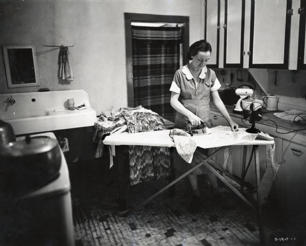 A woman irons clothing on an ironing board in a kitchen setting. She is standing in the middle of the room, surrounded by cabinets, a sink and mirror, and a stove.