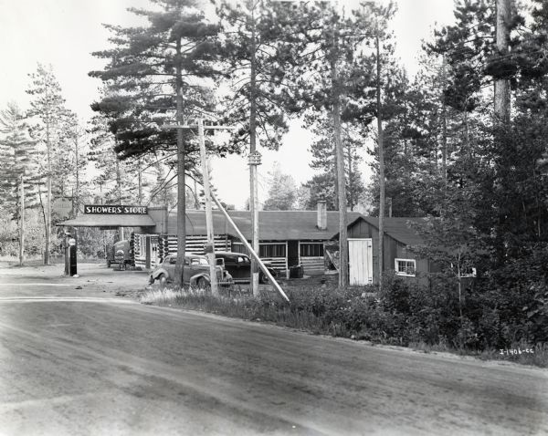 Exterior view from road of a log building with a sign reading: "Showers' Store." Automobiles are parked outside and a Shell filling pump stands near the store.