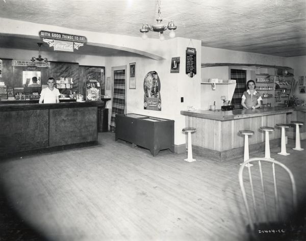 A man and a woman stand behind two separate counters inside a restaurant at Stone-Tavern. Signs on the walls advertise Coca-Cola, Schweppes, and read "With Good Things to Eat."