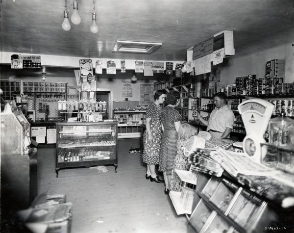 Two women and a girl stand near a counter inside a general store as a man shows them a product. A cat and a dog sleep on the floor in the background, and a sign reading: "Side Lake Post Office" hangs on the back wall.