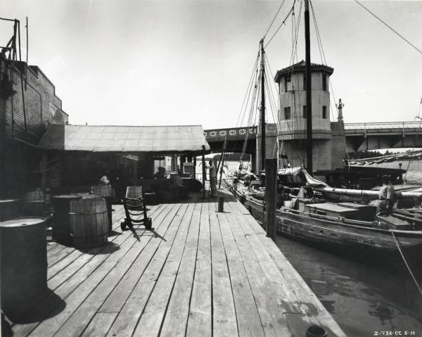 View of the dock area used by Mirabella Ice Company's wholesale and retail fish store. Men are standing and sitting near the receiving area, and boats are docked nearby. There is a bridge in the background.