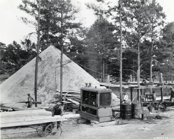 An International PD-80 diesel power unit used to power an A.B. Carroll Lumber Company sawmill. Several men are at work on the site; trees, and what appears to be a large sawdust pile are behind the workers.
