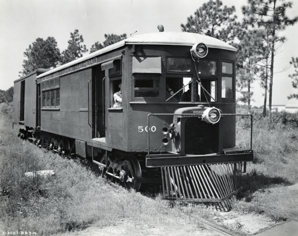 A motor car with an attached trailer drives down a railroad track. The car is operated by the Alabama & Florida Railroad Company and power is provided by an International PD-80 Diesel power unit.