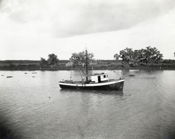 Three men on a boat called <i>Balbo</i> owned by Felice Golino of St. John's Shrimp Company. The boat was powered by an International PD-40 power unit and features an American flag. In the background is a shoreline.