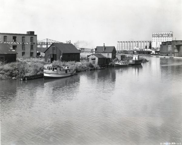 Elevated view of a boat owned by Julius Dettloff docked along the edge of a river. The boat was powered by an International PD-40 power unit; in the background are buildings with signs that read: "Stratton Grain Co." and "Atlas Storage Co."