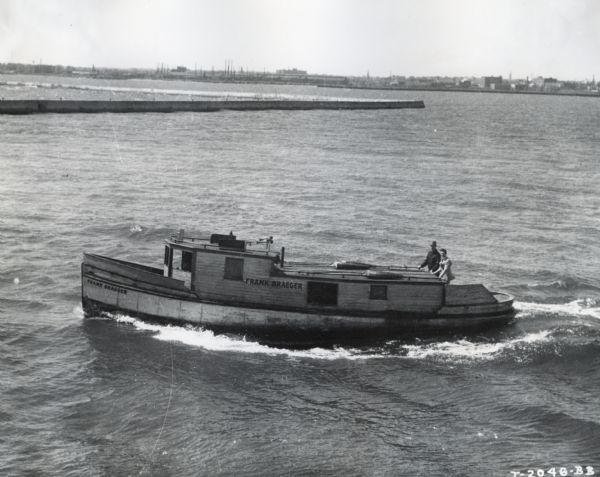 Two men stand to the rear of a 44-foot fishing boat marked <i>Frank Braeger</i>. The boat was owned by Julius Detlaff and powered by an International PD-80 power unit. In the background is what appears to be a jetty, and in the far background industrial buildings are along a shoreline. This boat is the new version of an earlier Frank Braeger fishing tug.