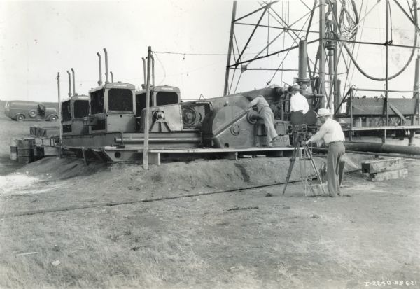 Four International PD-80 Diesel power units compounded on a drill rig owned by Taubert & McKee of Fort Worth, Texas, drilling a 4,500 foot hole two miles from Slidell. Two men stand near the power units while a man in the foreground sets up a motion picture camera to take a film of the scene.