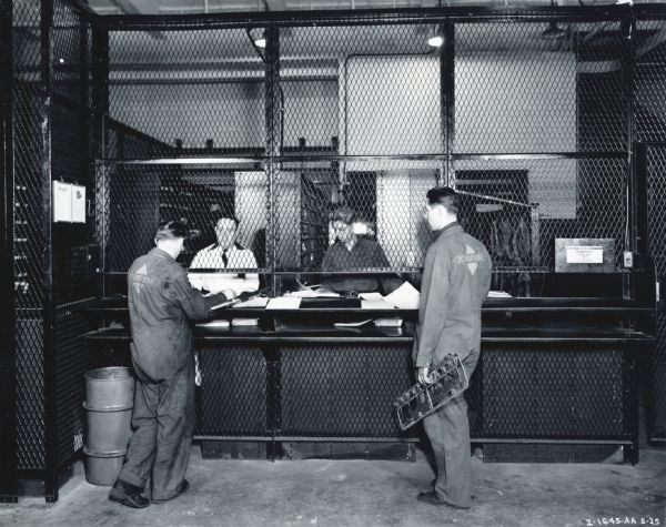 Two men wearing International jumpsuits wait to be assisted at the mechanics' parts counter at International's New York Manhattan truck branch. Two men work behind the counter, separated from the mechanics by a chain-link barrier.