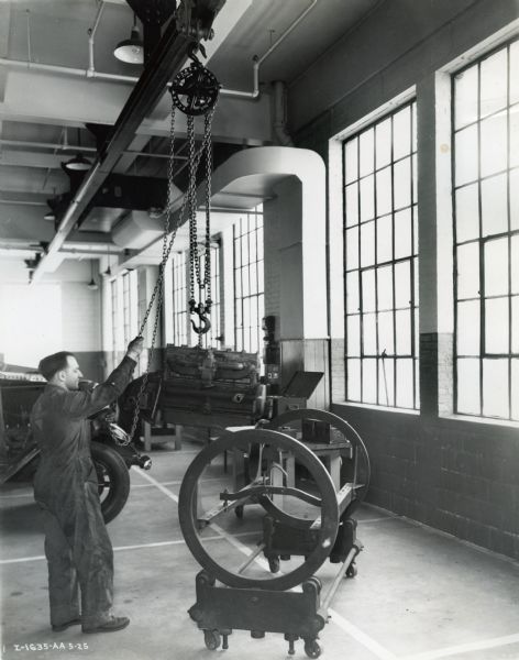 A man uses a chain pulley to lift what appears to be an engine in the service department on the second floor of International Harvester's New York truck branch.