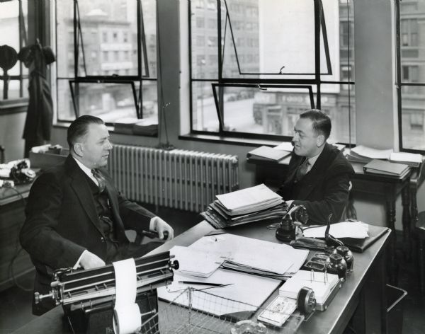 E.S. Dumble (left), office manager, sits behind a desk in an office at International Harvester's Manhattan truck branch while W.H. Lorenz, cashier, sits beside him.