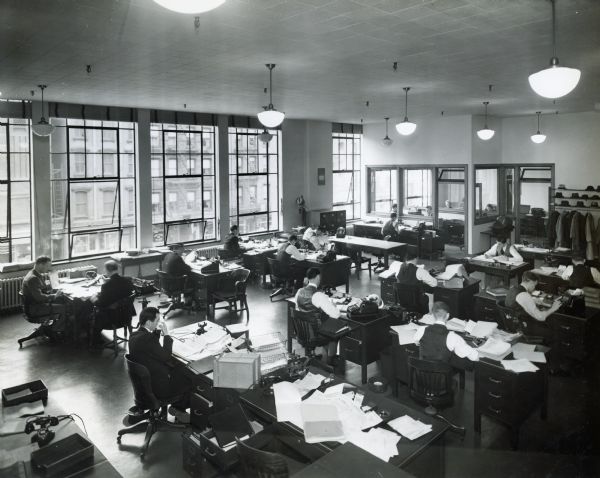 Elevated view of men sitting at desks near large windows in the general office of International Harvester's Manhattan truck branch. Additional men and women sit in an adjacent room behind the coat rack in the background.