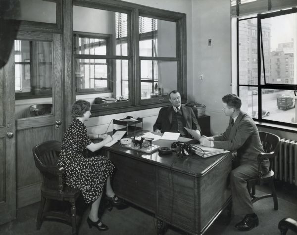 Olga M. Stankowitz, stenographer, E.O. Snider, credit and collection manager, and P.I. Brandjord, collection correspondent, sit around a desk in an office at International Harvester's Manhattan truck branch.