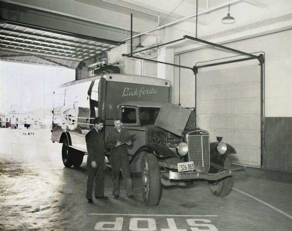 Two men stand near a truck in the receiving room of International Harvester's Manhattan truck branch. The truck bears the text: "Bickfords" and is parked near an open garage door.