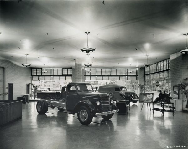 Three trucks parked in the showroom of International Harvester's Manhattan truck branch. Large show windows are in the background which look out on a street corner. An International engine sits on display to the right of the trucks along with a sign detailing its specifications.
