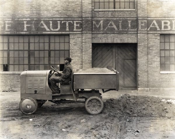 A man sits behind the wheel of a P-12 shop mule in a dirt lot in front of the Terre Haute Malleable & Manufacturing Company building.