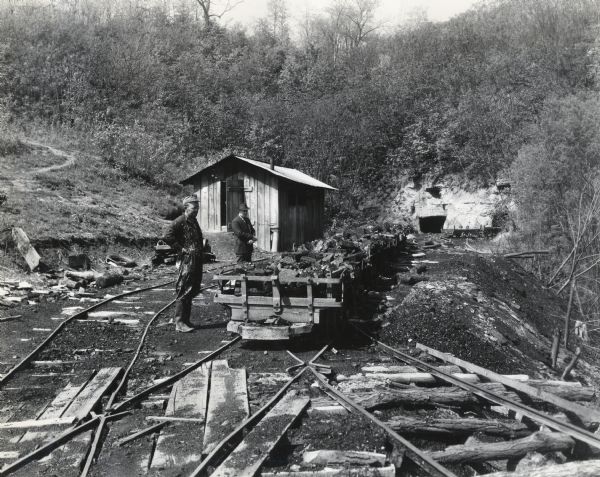 Two men standing next to mining cars near the opening of the Beam & Merritt mine as an International PA-50 power unit is installed.