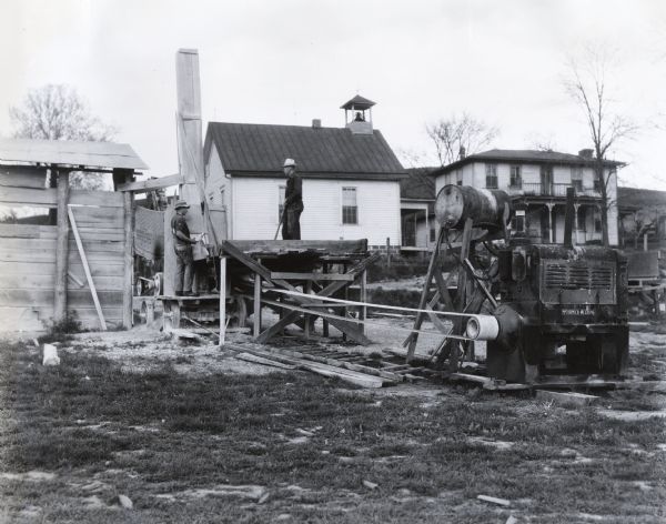 Men stand near a stone crusher powered by an International PD-40 power unit. The crusher was owned and operated by Coy Hawk.  Two buildings stand in the background; the building on the left appears to be a chapel or schoolhouse with a bell tower on the roof, and the building to the right appears to be a residence.