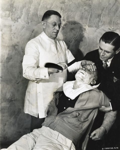 C.E. Jacquier, branch manager and defeated team captain of a tractor sales contest, is held by C.P. Wells, assistant branch manager, while a barber shaves his face.