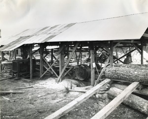 International P-40 power unit under a shed at a sawmill. Logs are piled near the sheltered work area, and a man works beside them.