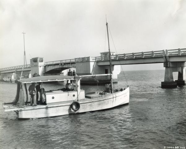 Men stand on <i>Jupiter</i>, a 35-foot shrimp boat owned by E.J. Chapman of Theodore, Alabama. The boat was powered by an International P-40 Marine engine. The boat is near a large bridge.