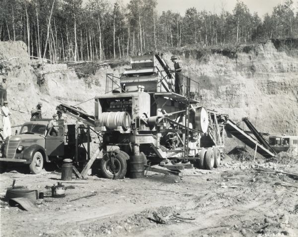 Men work with a Universal crusher, used to crush 400 yards of gravel in 8 hours. The crusher was powered by an International PA-100 power unit owned by E.F. Wick; an International truck is to the left of the crusher.