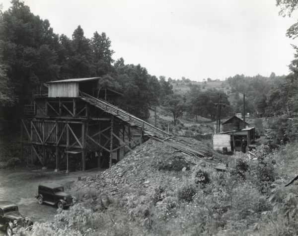 Buildings owned by the Peacock Coal Company. A man is standing beside a small wooden structure on the side of a hill. There is a ramp angling up and away from the side of the hill to a muti-storied structure on the left. Two automobiles are parked nearby. Operating power for the mine was provided by an International PA-100 power unit.