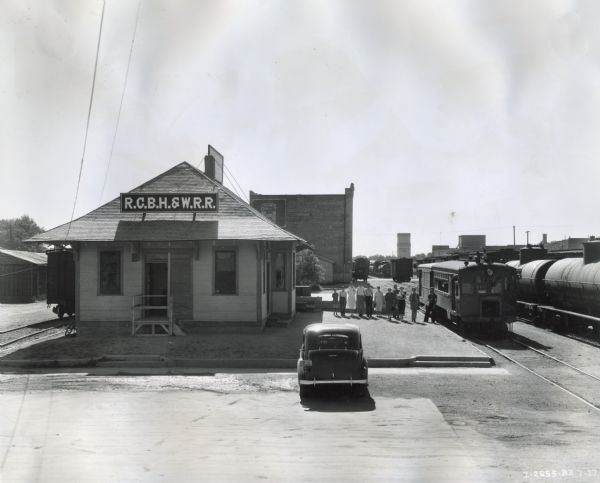 View across road of a row of men and women standing beside a rail car powered with an International PA-100 power unit. To the right of the car is another train and to the left are the depot building and a parked automobile.