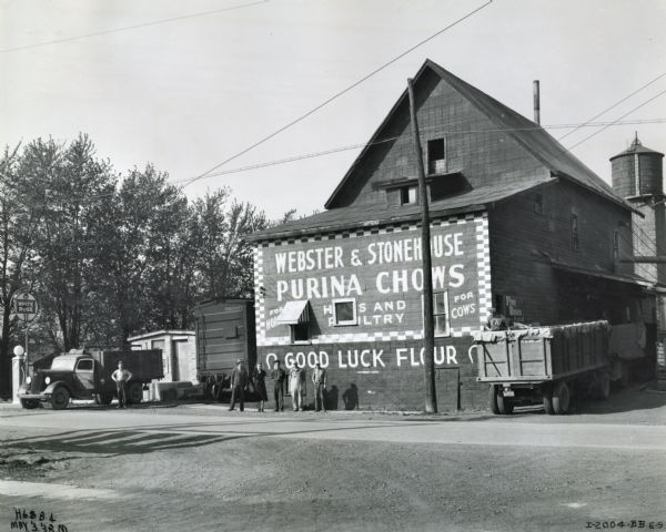 View from across road of six men and one woman standing outside the Webster and Stonehouse flour mill. Another man stands in the back of a truck to the right of the building.